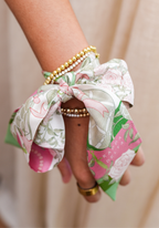 Silk scarflette with pink, green, and white floral pattern tied around wrist with gold and pearl bracelets.