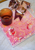 Pink acrylic tray with gold scalloped edges, floral and animal print accents, amber glass, and silk scarf with giraffe design.