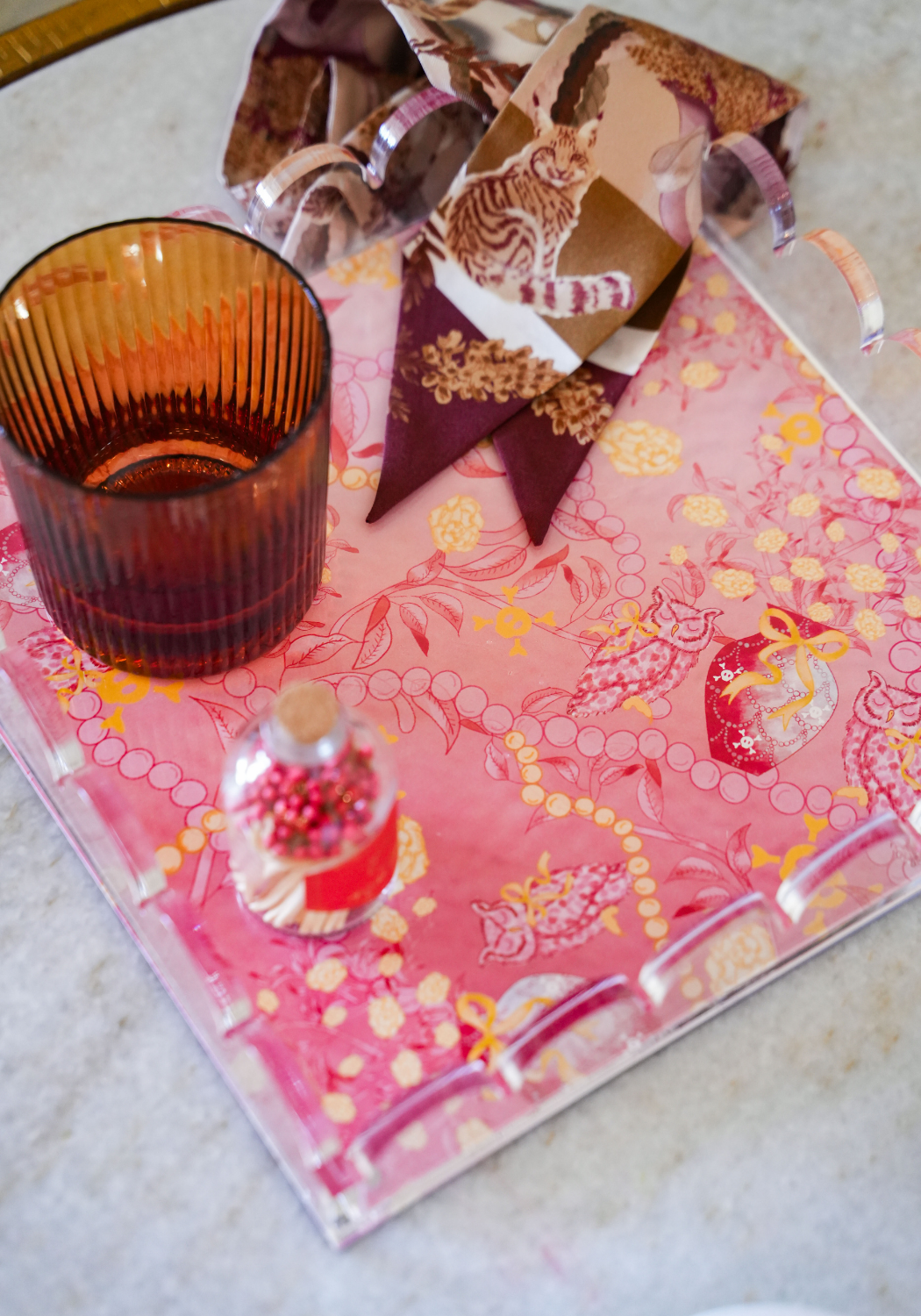 Pink acrylic tray with gold scalloped edges, floral and animal print accents, amber glass, and silk scarf with giraffe design.