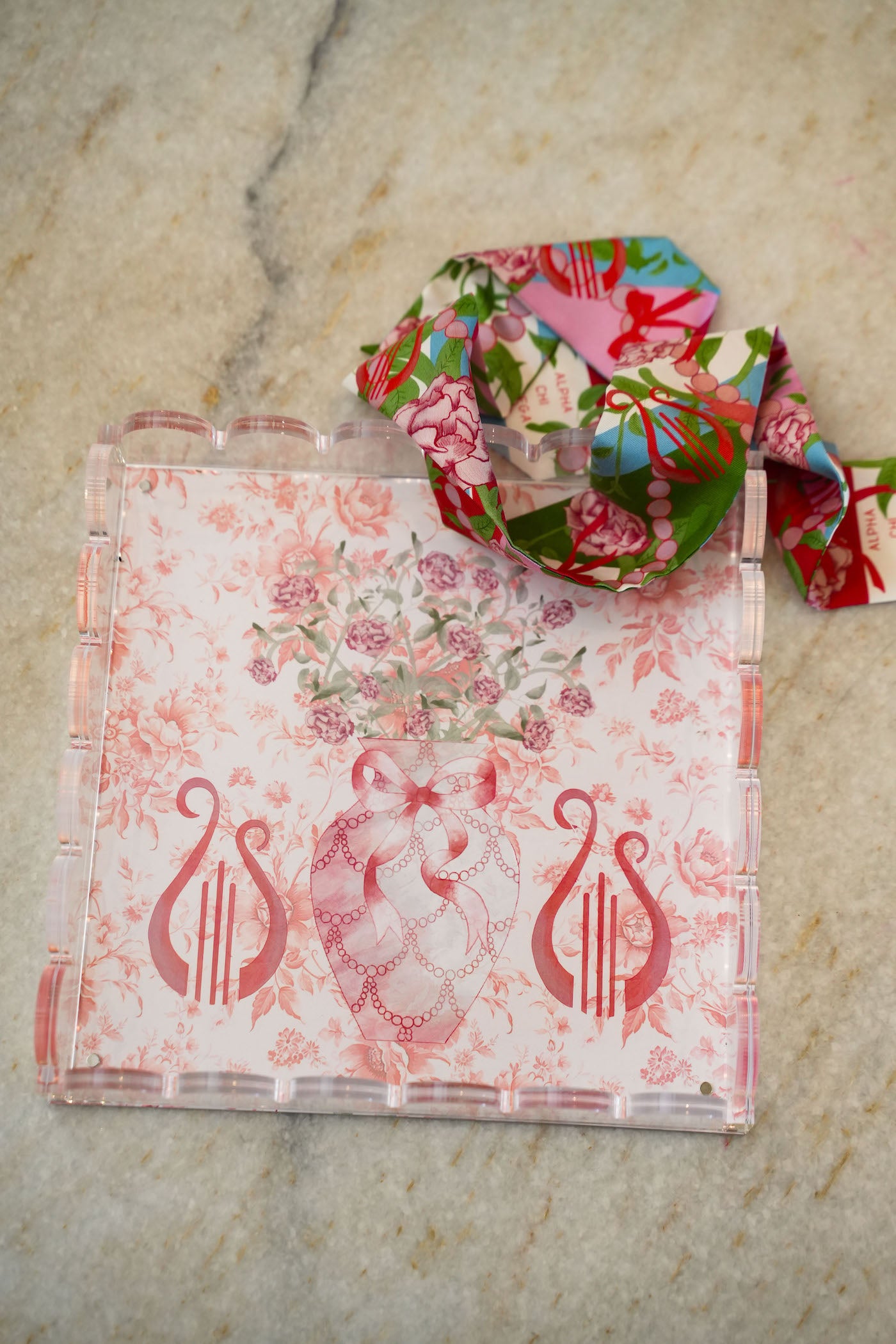 Clear acrylic tray with scalloped edges, pink floral and lyre designs, and a matching floral ribbon looped on top.