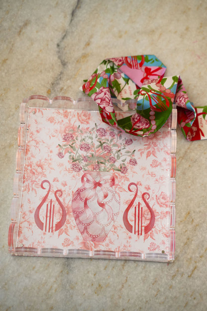 Clear acrylic tray with scalloped edges, pink floral and lyre designs, and a matching floral ribbon looped on top.