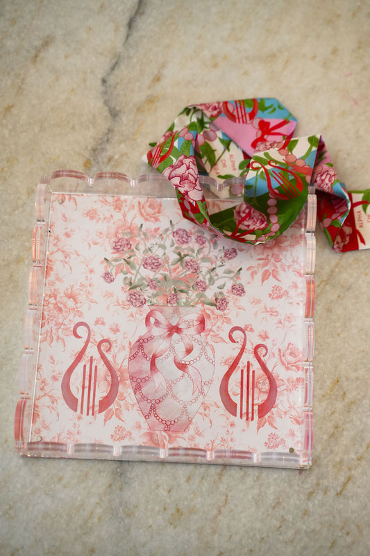 Clear acrylic tray with scalloped edges, pink floral and lyre designs, and a matching floral ribbon looped on top.