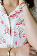 White button-up pajama top with pink Alpha Chi Omega crest and floral print, front pocket, and gold bracelets on wrist.
