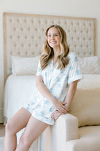 Light blue and white tie-dye short-sleeve pajama top and matching shorts on a smiling woman sitting on a beige upholstered.