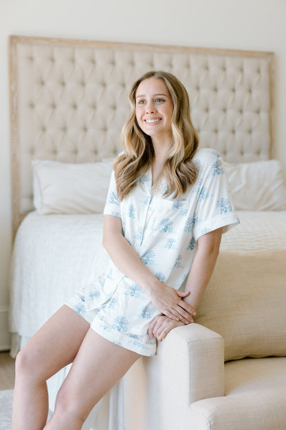 Light blue and white tie-dye short-sleeve pajama top and matching shorts on a smiling woman sitting on a beige upholstered.