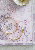 Clear acrylic tray with scalloped edges featuring a pink floral and teddy bear design and gold hoop earrings placed on top.
