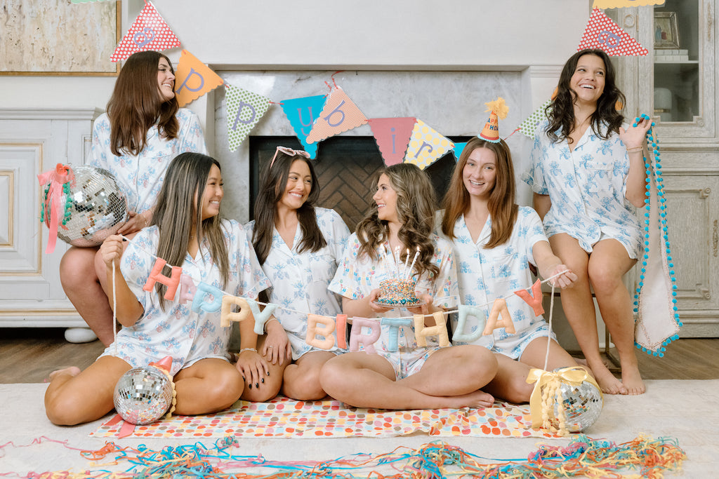 Six women wearing colorful birthday pajama shorts sets with party hats and birthday decorations indoors.