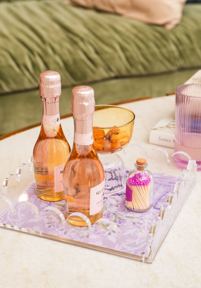 Clear acrylic tray with a purple floral base holding two small rose gold bottles, a bowl of orange snacks, and a cupcake.